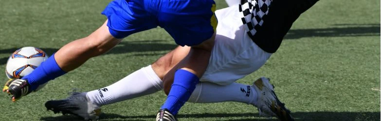 Nahaufnahme der Hände eines Tennisspielers mit einem weißen Schweißband, der einen gelben Ball an die Bespannung eines schwarz-blauen Tennisschlägers hält. Der Hintergrund zeigt einen orangefarbenen Sandplatz mit einer unscharfen weißen Linie.